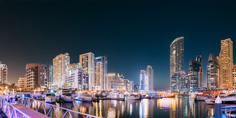 Obraz premium Dubai Marina Port, UAE, United Arab Emirates - Beautiful Night View Of Dubai Marina Towers. Yachts Moored Near City Pier, Jetty In Evening Night Illuminations. Panorama, Panoramic View.