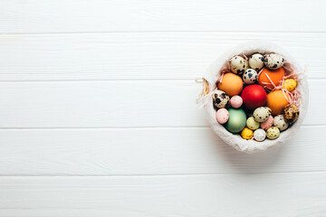 Colored eggs of different sizes in a straw basket on a wooden white background. Symbol of the Easter holiday. Easter background. Flat lay