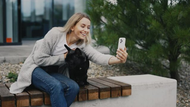 Beautiful Smiled Caucasian Woman is Sitting on the Bench with Her Lovely Black French Bulldog and Doing Selfie, Making Memories with Her Puppy. Technology and Dogs Friendship Concept