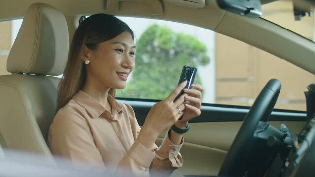 Side view of elegant Asian businesswoman sitting on driver seat in car, holding hand on steering wheel and having talk on mobile phone