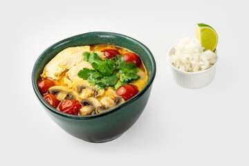 Spicy creamy soup with chicken, cherry tomato, cilantro leaves, mushrooms and little bowl of rice with slice of lime on light background. Asian Thai food