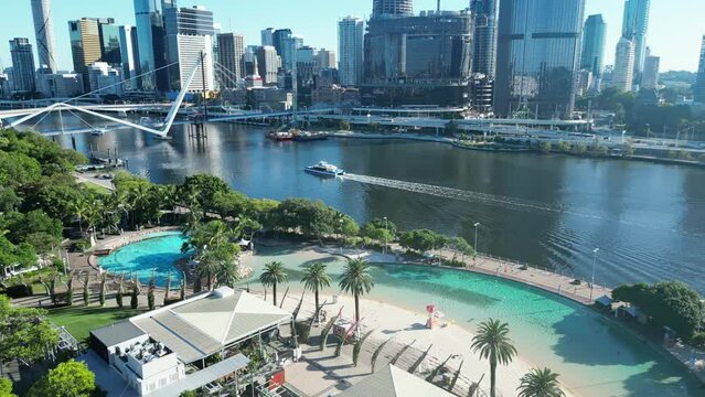 Drone shot of Brisbane City's South Bank Beach and Parklands. Slow camera movement, Shot at sunrise. Camera tracking a City Cat Boat while it's crossing Brisbane River.