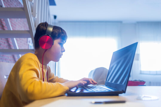 Side View Of Boy Doing Homework Using Headphones And Laptop Computer At Home