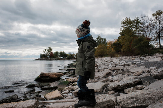 Side view of boy wearing warm clothing looking at sea while standing on rocks against stormy clouds