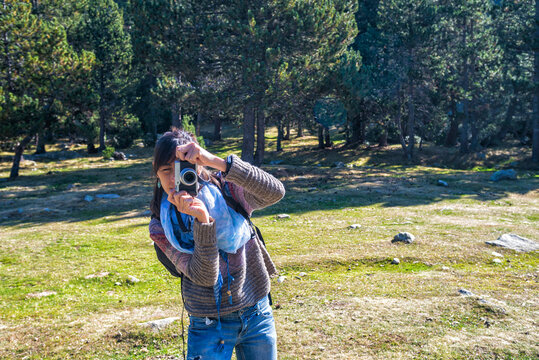 Female hiker photographing with camera while standing against trees in forest during sunny day