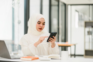 A portrait of an Asian Muslim girl wearing a hijab, using her smartphone at home, messaging or browsing social networks while sitting on a sofa in the living room.