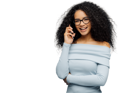 Glad African American female enjoys telephone conversation, keeps one hand over waist, uses modern technologies, wears spectacles and light blue jumper, stands in studio, blank space for your slogan