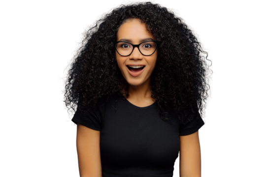 Pleasantly surprised dark skinned woman keeps jaw dropped, gazes with interest, has curly hair, wears black t shirt, models against blue wall. Afro American lady gazes with shock, has bated breath