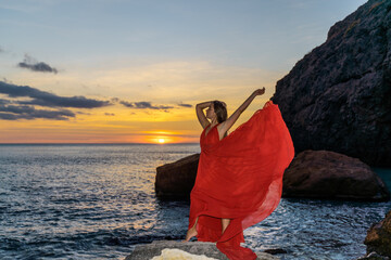 Woman in a red flying dress on the ocean or on the sea beach against the backdrop of the sunset sky.