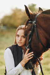 Fototapeta premium Young beautiful female jockey strokes and hugs the horse's head and prepares for the competition. Jumping training in the meadow in summer evening.