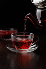 Hot tea is poured from a glass teapot into a cup on a black background. Selective focus.