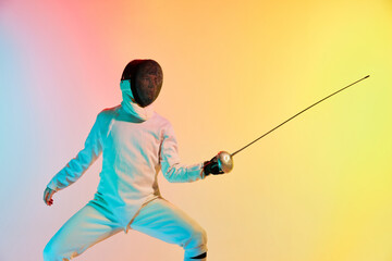 Young man, male fencer with sword practicing in fencing over gradient pink-yellow background in neon light. Sportsman shows fencing technique
