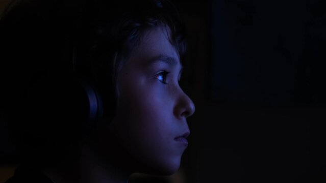 young boy is seen sitting in a dimly lit room, his face illuminated by the flickering glow of a television screen. child is watching TV late at night.
