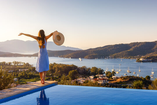 A happy holiday woman in a dress stands at the swimming pool and enjoys the summer sunset behind the mediterranean sea