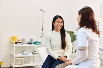 A happy female Asian patient talking with a female doctor in the examination room.