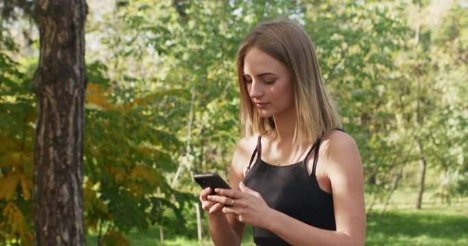 Young woman wearing sportsuit walks in park scrolling social media on smartphone. Short-haired lady checks running progress on phone app