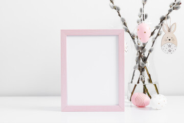 Easter holiday composition. Empty pink photo frame mockup, vase with willow branches, easter eggs, bunny on white table background. Minimal conceptEaster. Front view