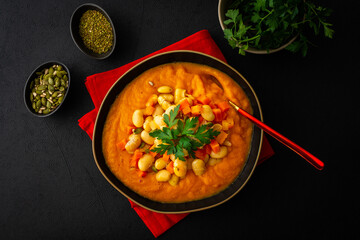 Healthy lunches, tomato and carrot puree soup, beans with pumpkin seeds, healthy tasty legumes, top view of a plate of soup on a black background