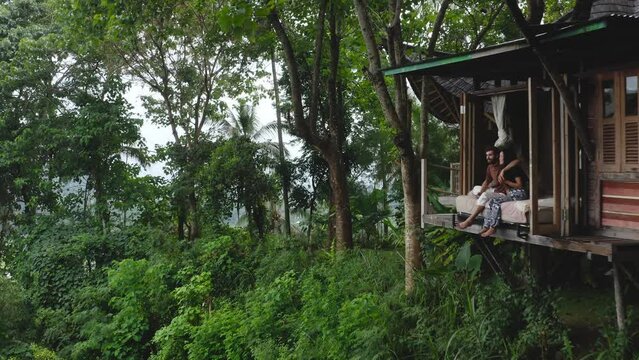 Romantic couple sit by treehouse cabin in forest in Indonesia, aerial pull-out