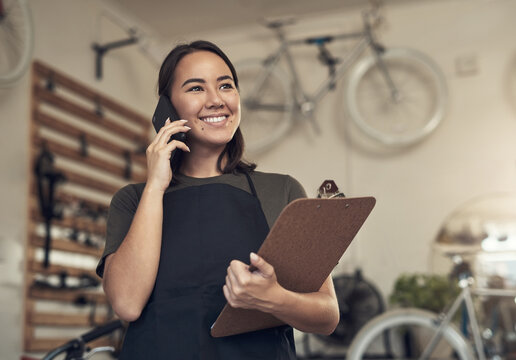 You can come through to collect your bike. Shot of an attractive young woman standing alone in her bicycle shop and using her cellphone while holding a clipboard. - Powered by Adobe