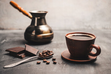 Black coffee drink in a clay cup, turkish jezve coffee pot, chocolate pieces and coffee beans in a spoon on dark background