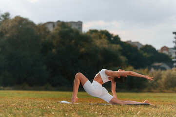 Side view. Doing yoga. Young caucasian woman with slim body shape is in the fitness clothes outdoors