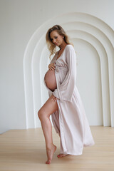 Beautiful pregnant woman in white retro underwear and gown standing near bath. Young blond lady in spacious room with lamps and window. Big pregnant naked belly. Pretty woman waiting child.