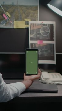 Vertical video: Law officer holding mobile pohne with greenscreen, young detective looking at blank copyspace display. Female detective in incident room using chroma key mockup template in archive.