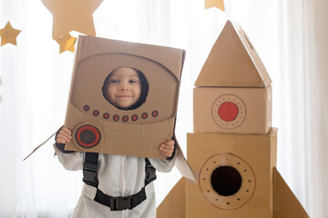 Sweet toddler boy, dressed as an astronaut, playing at home with cardboard rocket and handmade...
