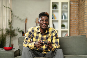 Young happy African guy playing video game at home. Man holding joystick and playing video games