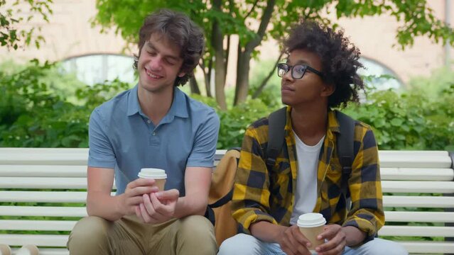 Multiethnic college students sitting on bench relaxing after classes drinking coffee