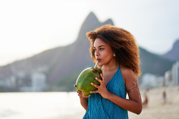 brazilian black young woman drinking coconut water on the beach at sunset in Ipanema, Brazil	