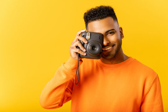 Smile. Cheerful glad Indian guy in casual t-shirt taking photo of you, holding instant photo camera and looking at the camera isolated on yellow, happy guy smiling with wide toothy smile. Studio shot