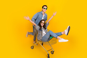 Full size photo of two carefree cheerful people sit ride market trolley isolated on yellow color background - obrazy, fototapety, plakaty