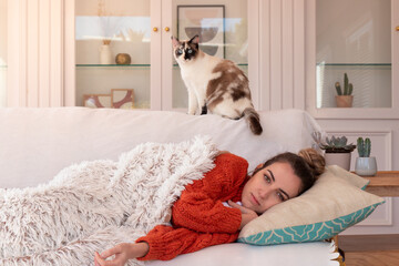 Sick woman with paper tissue lying with flu on comfortable couch near white and brown cat in light living room