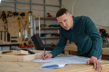 Portrait of a carpenter in a plaid shirt draws a workshop blueprint. 