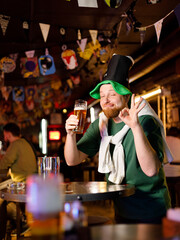 a real ginger guy in a leprechaun hat for st. patrick's day holds a glass of beer and shows ok