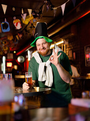 a real ginger guy in a leprechaun hat for st. patrick's day holds a glass of beer and shows ok
