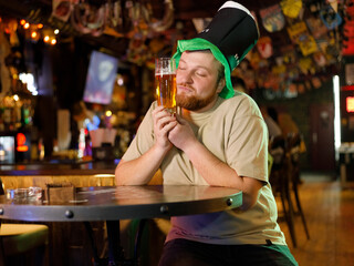 a guy with a red beard in a leprechaun hat hugs a glass of beer