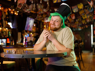 a guy with a red beard in a leprechaun hat hugs a glass of beer