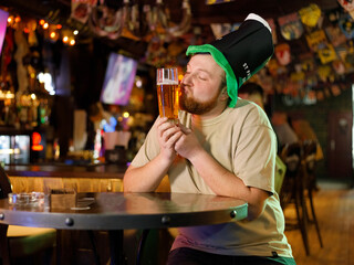 a guy with a red beard in a leprechaun hat kisses a glass of beer