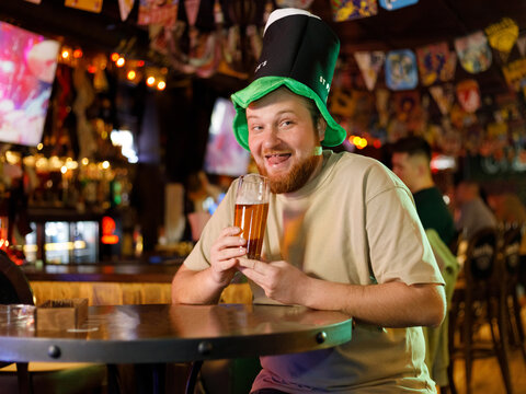 a guy with a red beard in a leprechaun hat fools around with beer