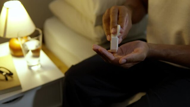 Unrecognizable person taking pills at night. Black mand sitting in bed holding medication and glass of water before sleep. 