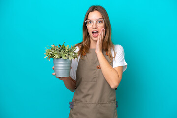 Young caucasian gardener woman holding a plant isolated on blue background with surprise and...
