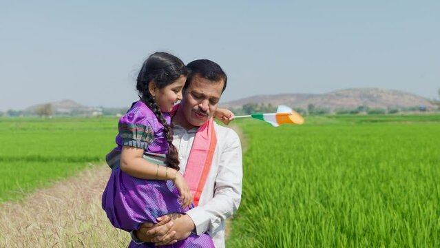 tracking shot of Happy village father carrying daughter with indian flag by walking near green farmland during independence day - concept of democracy, patriotism and relationship bonding.