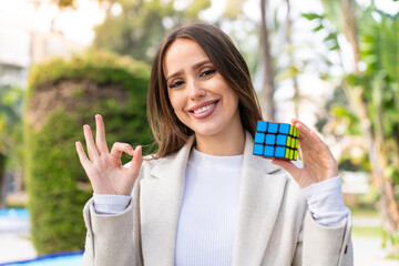 Young pretty woman holding a three dimensional puzzle cube at outdoors showing ok sign with fingers