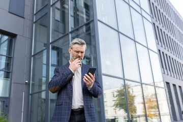 Fototapeta premium An older gray-haired man in a suit and glasses stands outside an office, a skyscraper. Worriedly looks at the phone, received a message, reads, calls.
