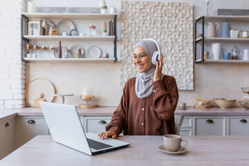 Young beautiful Muslim student woman in hijab sitting at home in kitchen wearing headphones and...