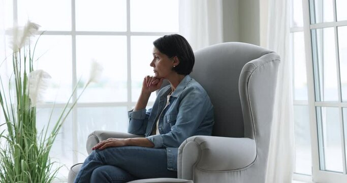 Sad depressed mature woman sitting in armchair at home, looking at window away in deep thoughts, thinking over elderly age health problems, feeling melancholy, loneliness, apathy. Side view