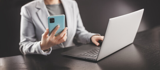 Woman in the office holding in her hands mobile phone and typing on the laptop.